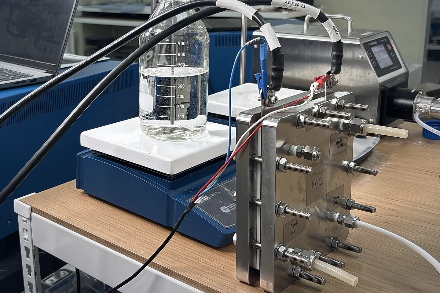 Lab bench with a single-cell test fixture connected to an electrolyte bottle and power equipment